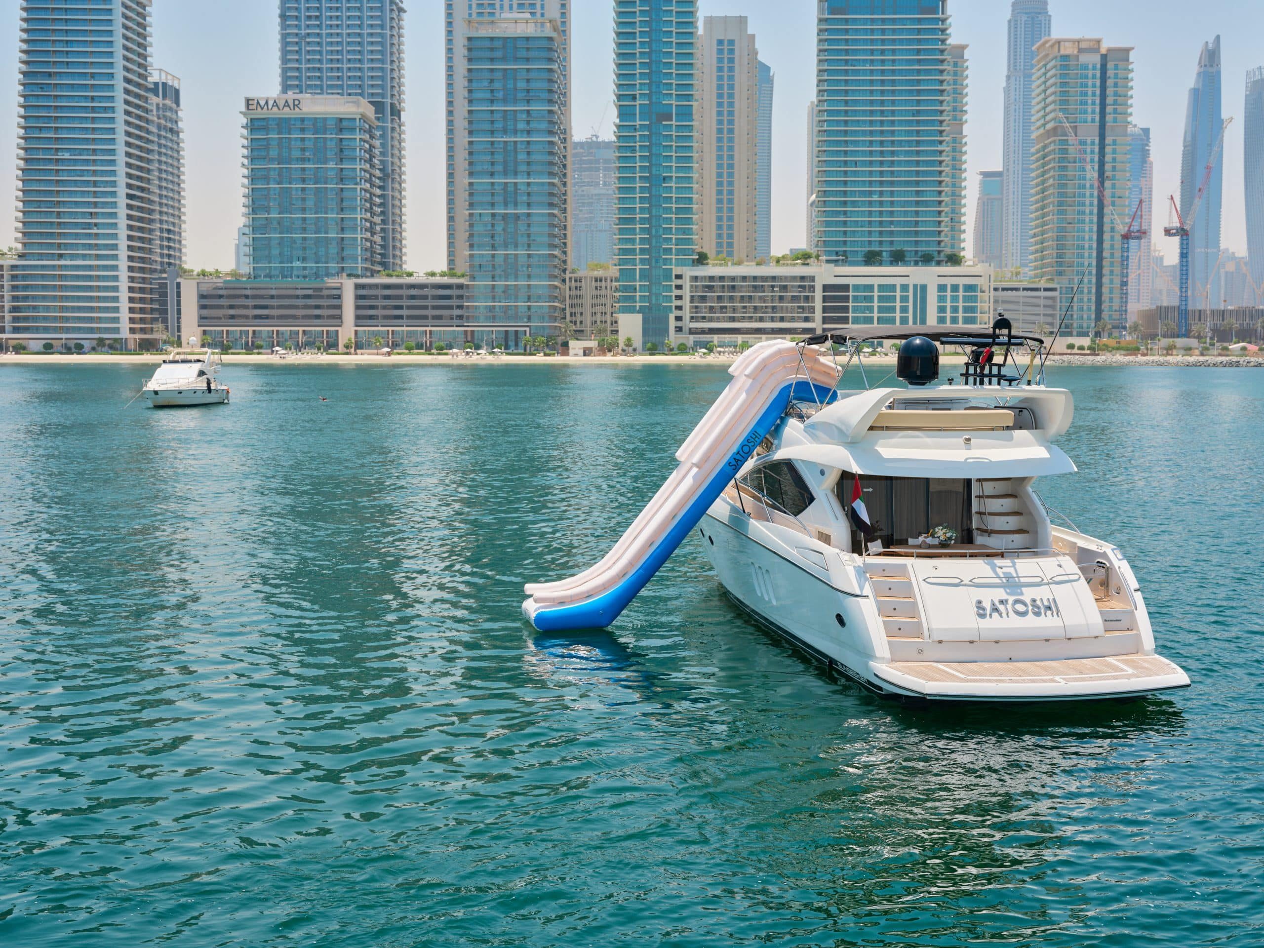 White yacht with an inflatable slide docked on calm water, with modern high-rise buildings and a city skyline in the background.