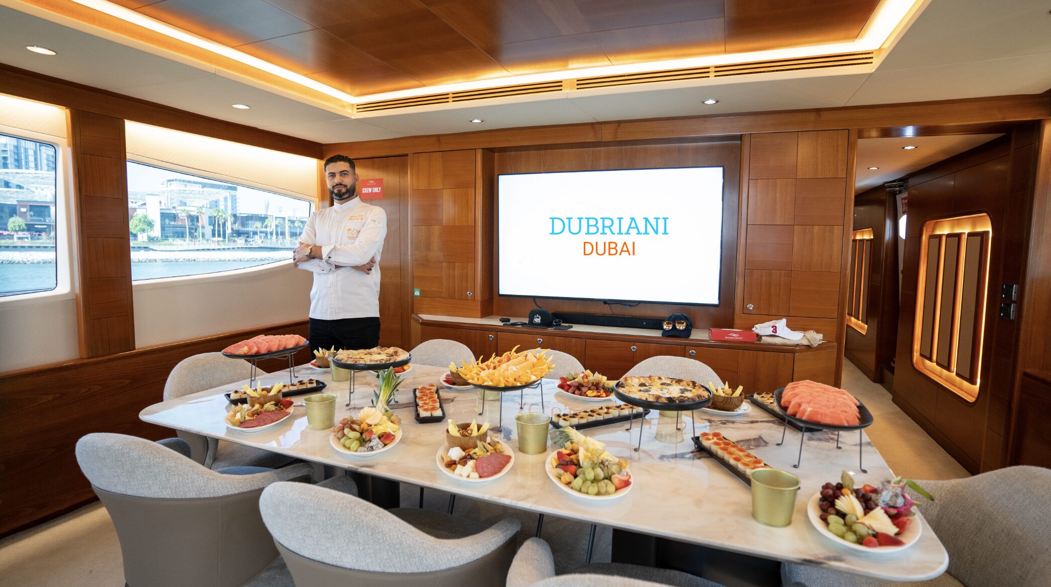 Chef standing beside a long buffet of fresh platters laid out in a wood-panelled yacht dining salon