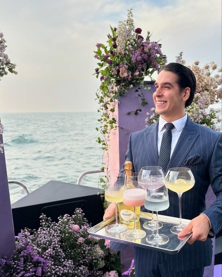 Server in a pinstripe suit carrying a tray of champagne glasses beside floral arrangements on deck at sea