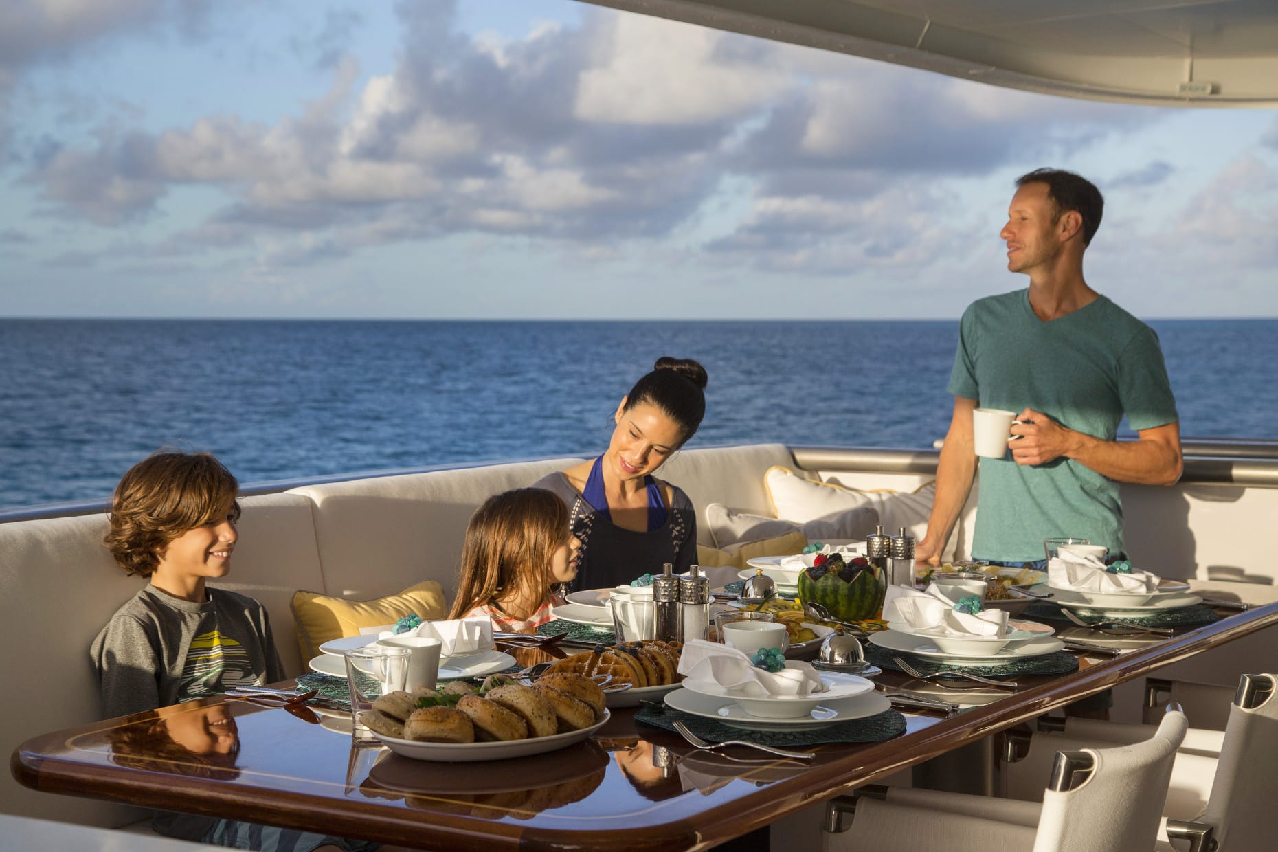 Family sharing a meal at a laid table on a yacht's aft deck with open sea behind