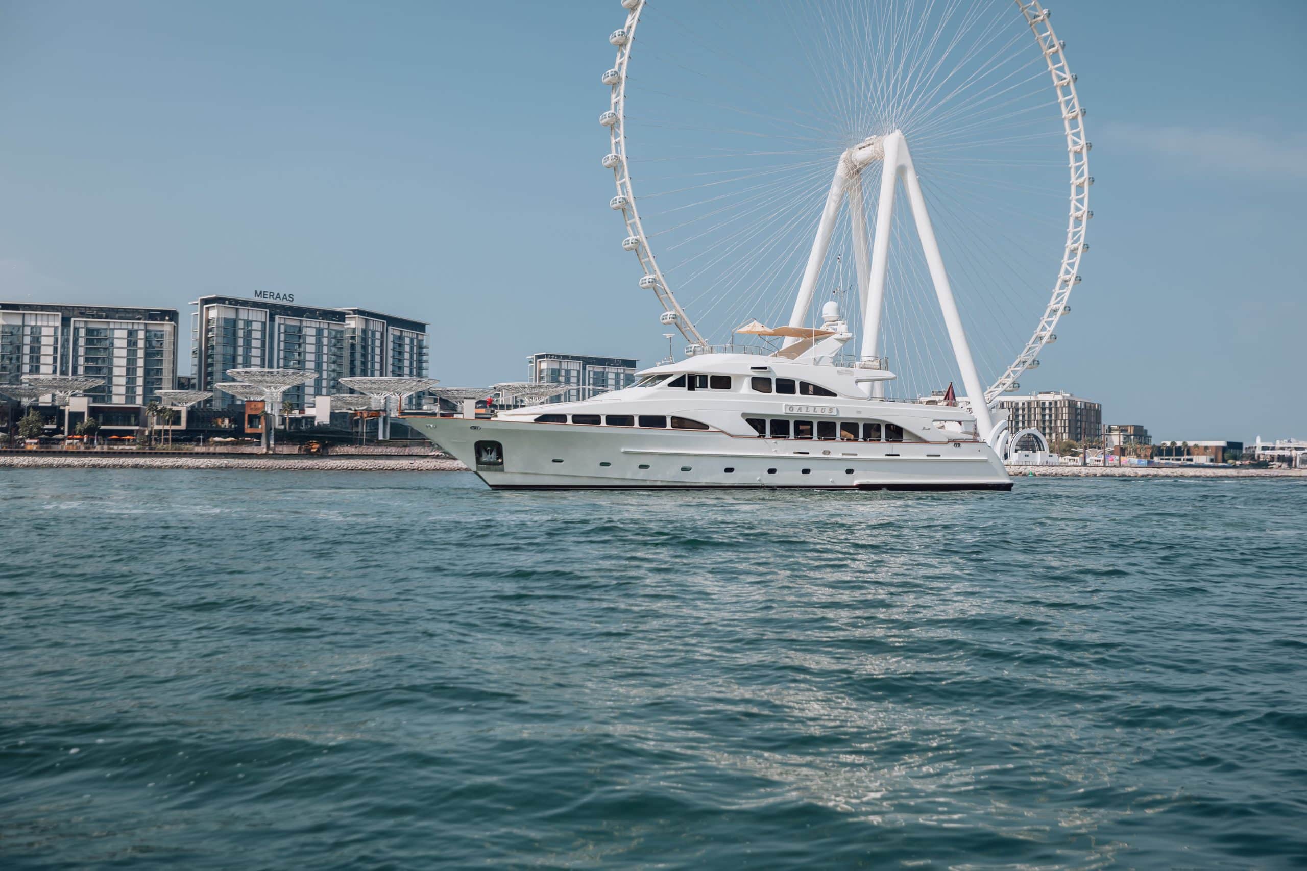 White luxury yacht sailing across turquoise waters with Ain Dubai and modern waterfront buildings in Dubai under a clear blue sky.