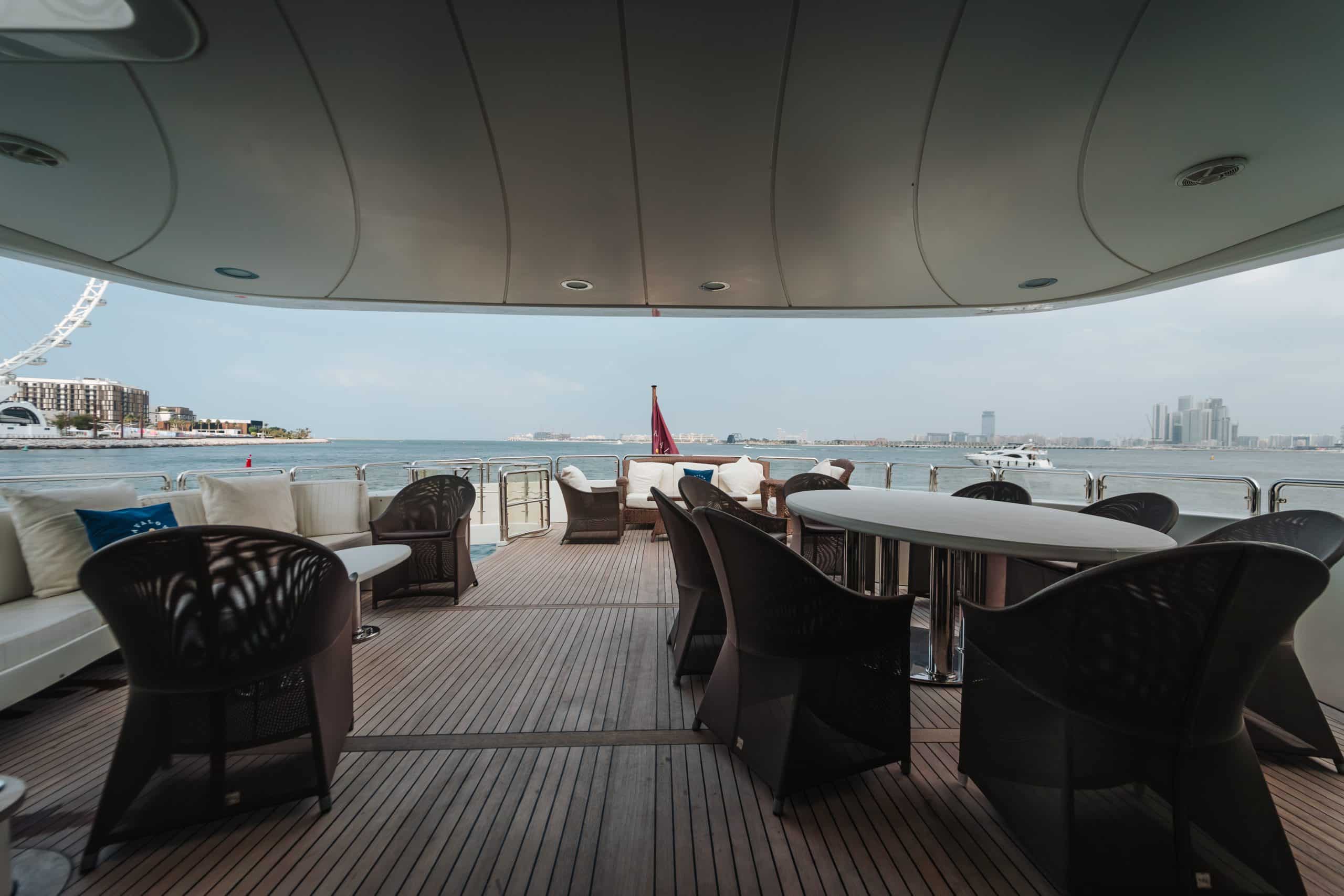 Outdoor seating area on the shaded upper deck of a luxury yacht, with tables and chairs overlooking calm water, a marina, and a modern city skyline in the distance.