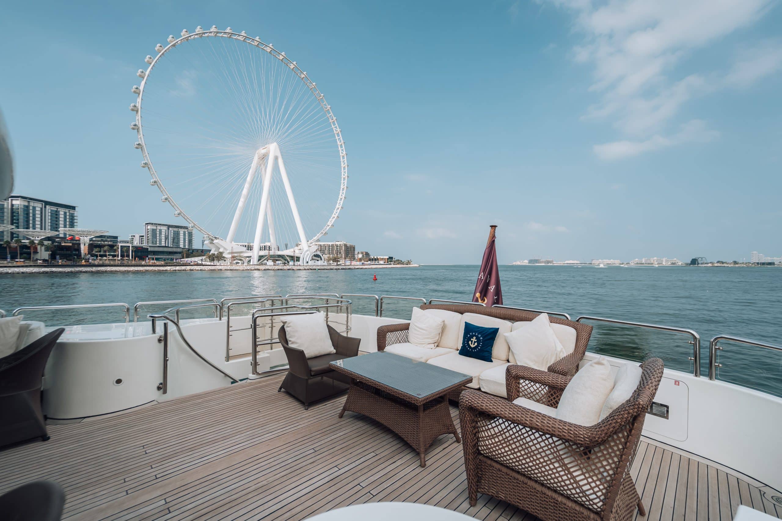 Lounge seating on the open deck of a luxury yacht, with wicker chairs and a sofa facing calm water and a large observation wheel and modern waterfront buildings in the background.