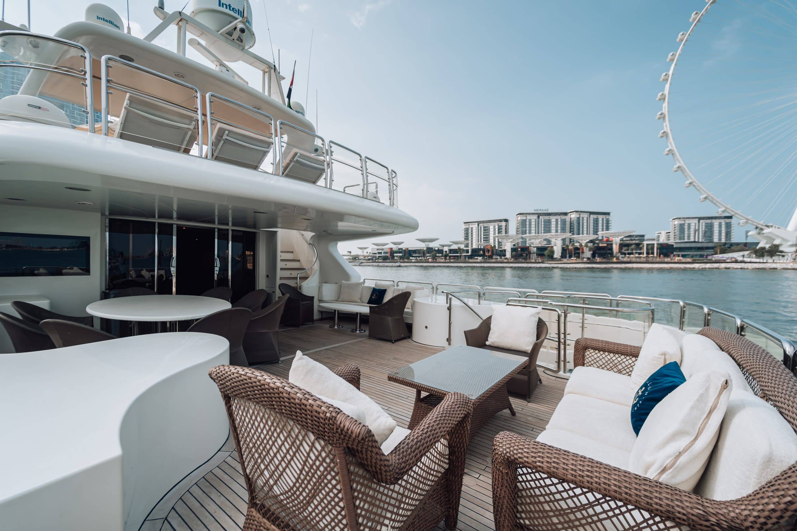 Shaded seating and dining area on the aft deck of a luxury yacht, with wicker lounge furniture, round tables, and a waterfront skyline with a large observation wheel in the background.
