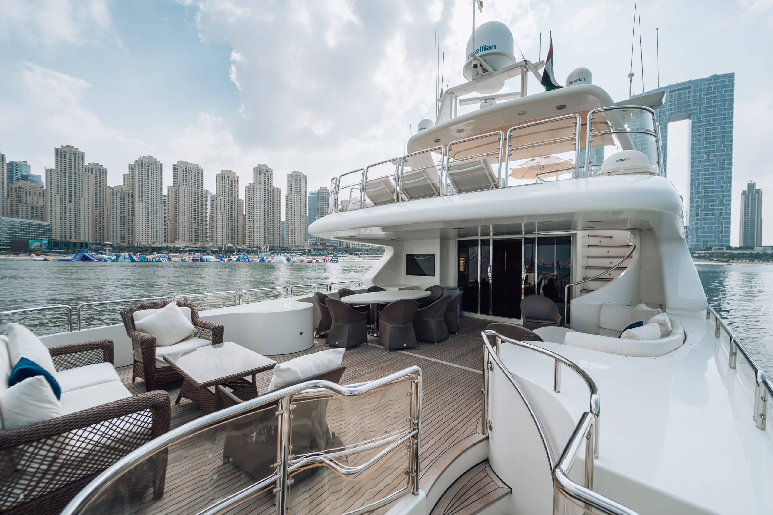 Spacious aft deck of a luxury yacht with wicker lounge seating and dining tables, overlooking calm water and a dense modern waterfront skyline with high-rise towers in the background.