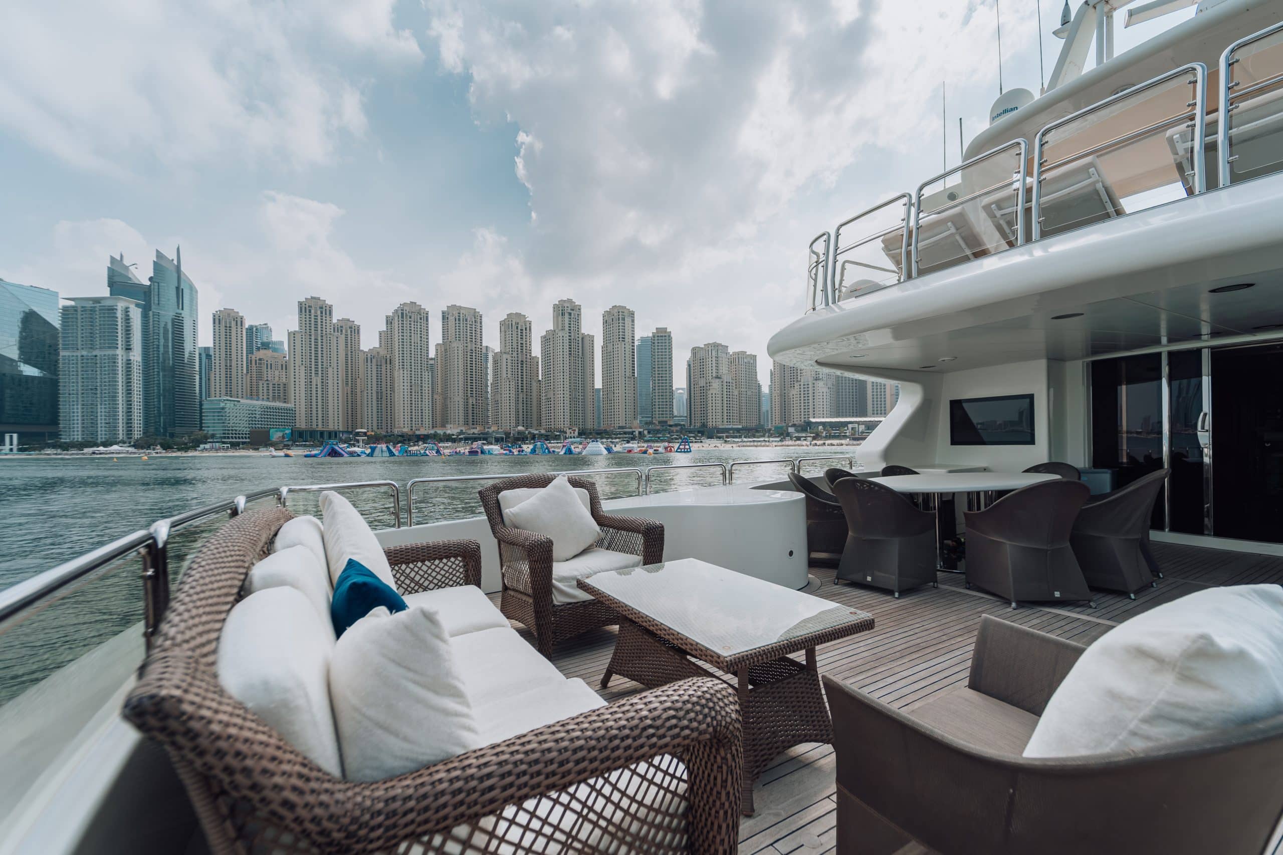 Wicker lounge seating and a low table on the open aft deck of a luxury yacht, overlooking calm water and a dense skyline of modern high-rise buildings across the marina.