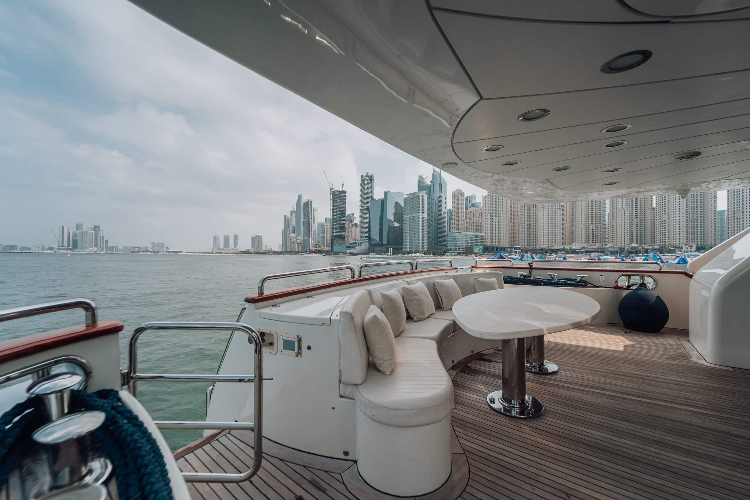 Curved built-in sofa and round dining table on the shaded rear deck of a luxury yacht, overlooking calm water and a modern city skyline across the marina.