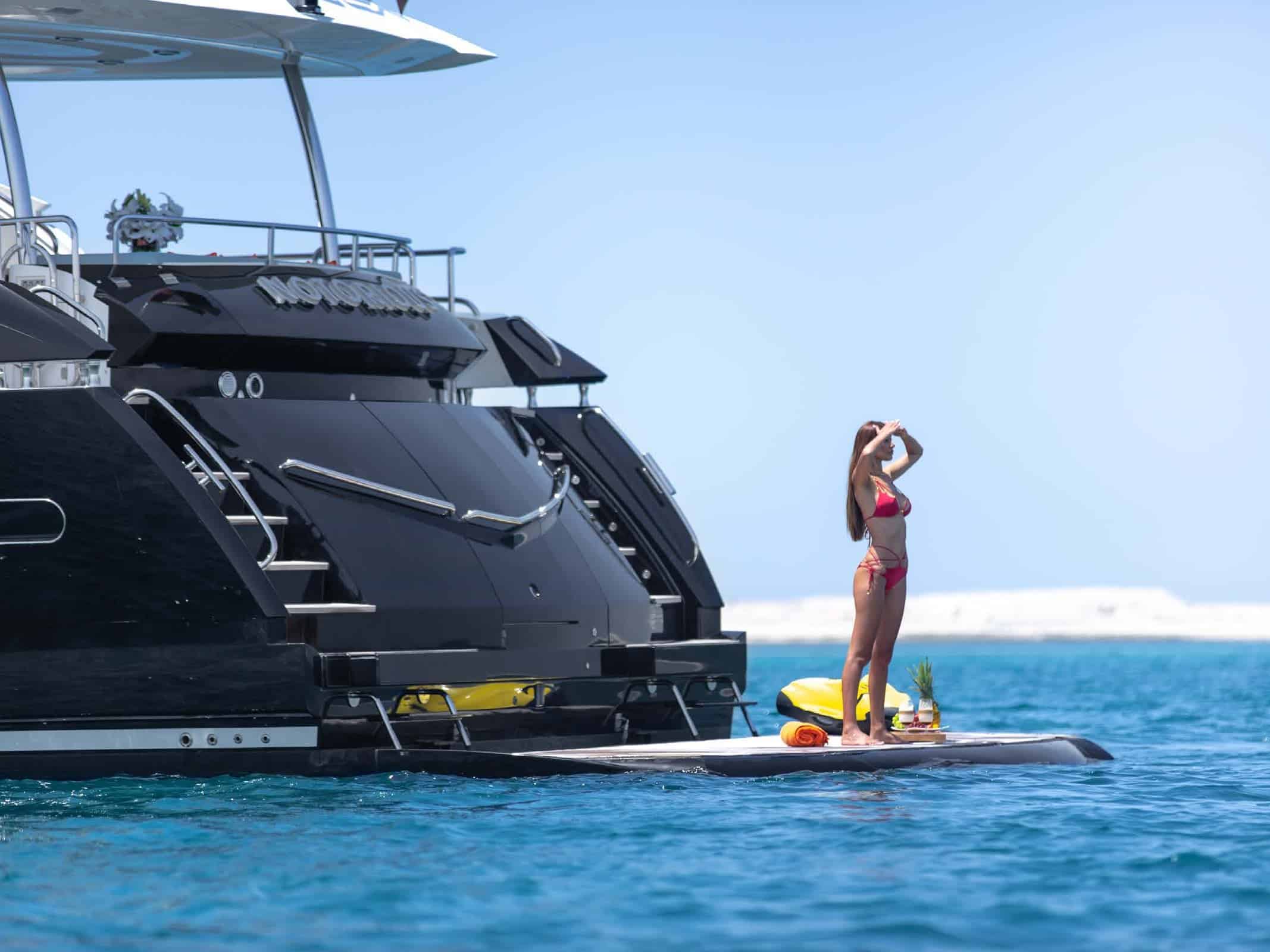 Notorious luxury yacht with guest standing on paddleboard beside swim platform