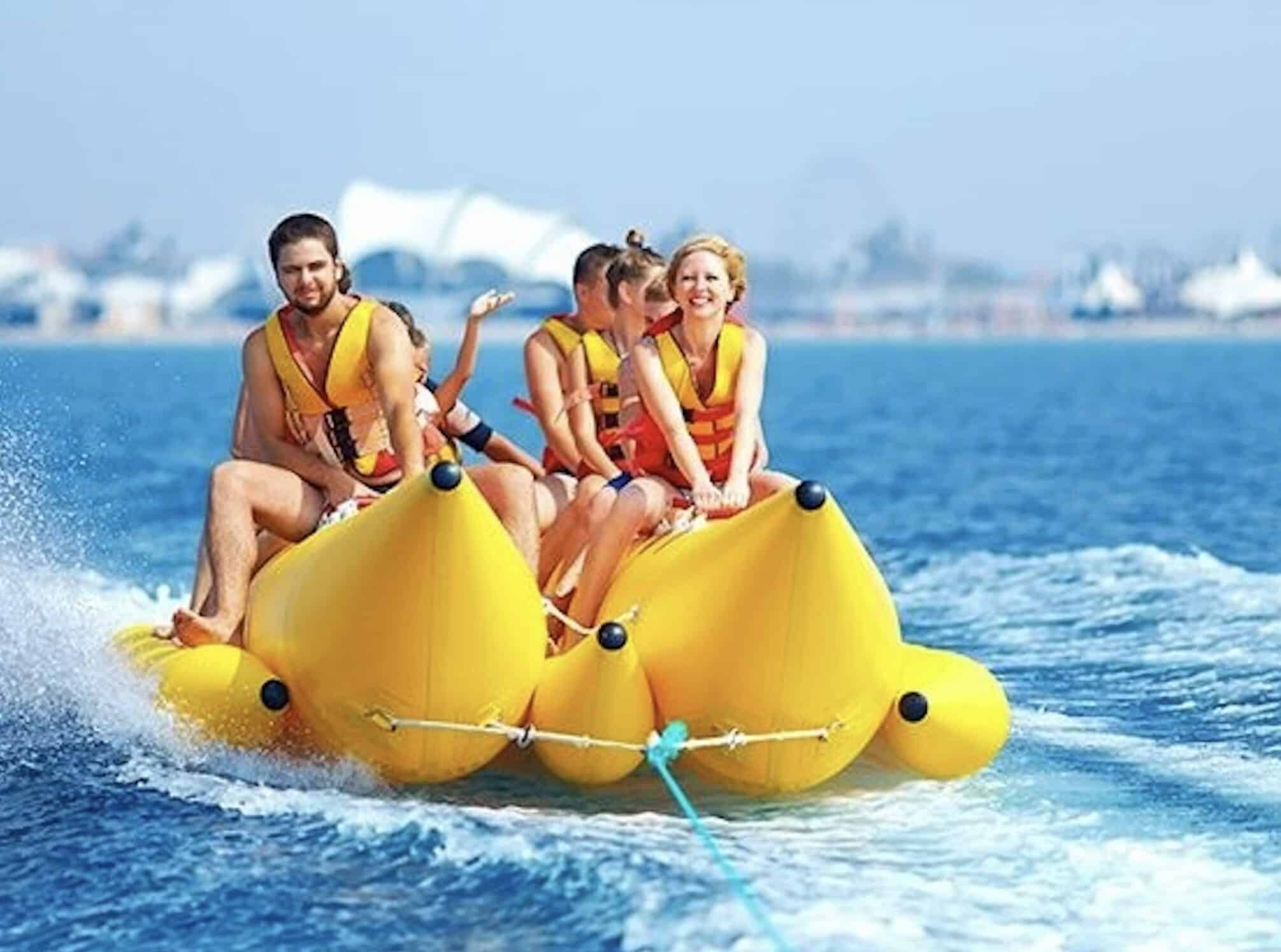 Group of five in orange life jackets laughing on a yellow banana boat towed across blue water