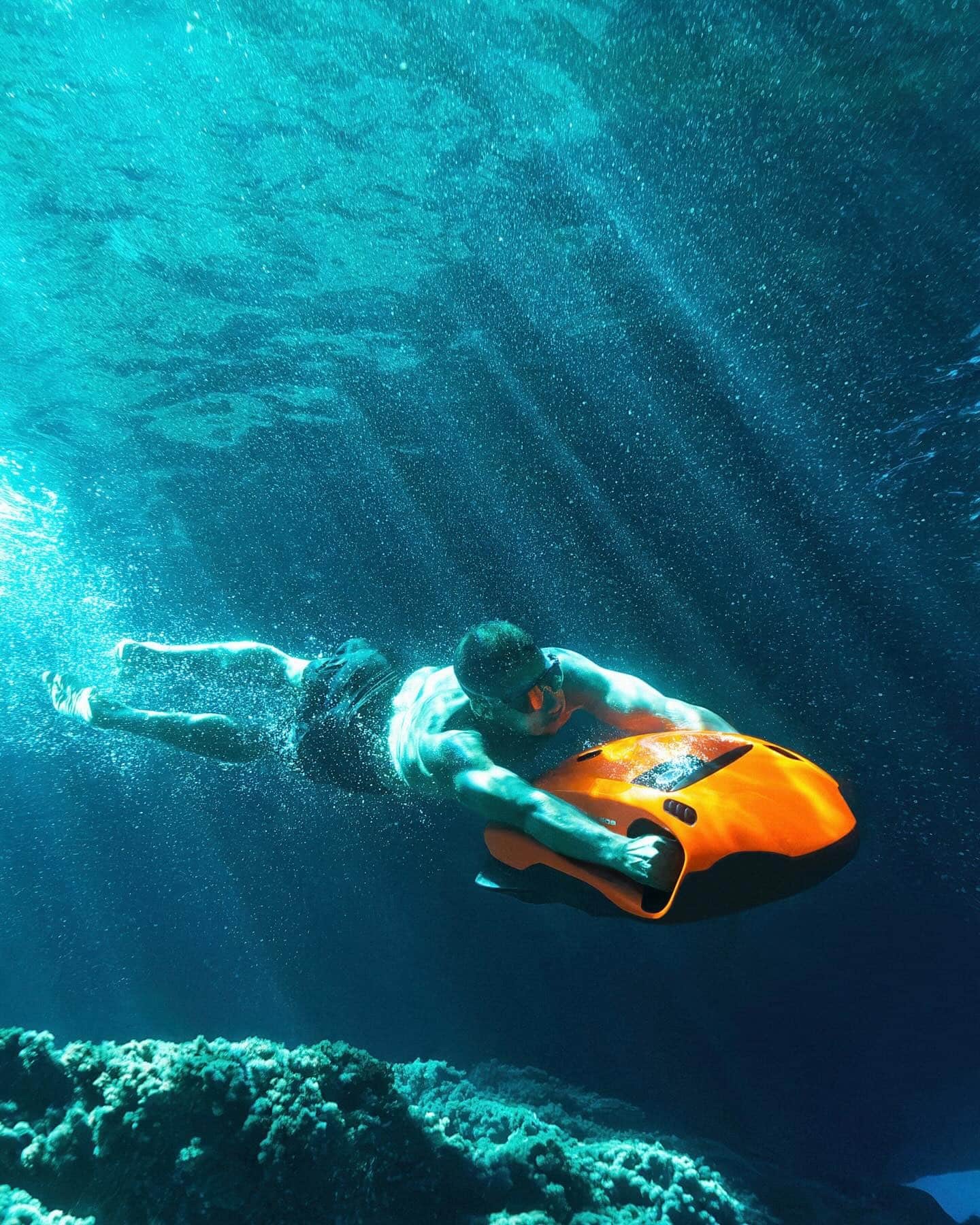 Person riding an orange Seabob underwater with sunlight streaming through the ocean