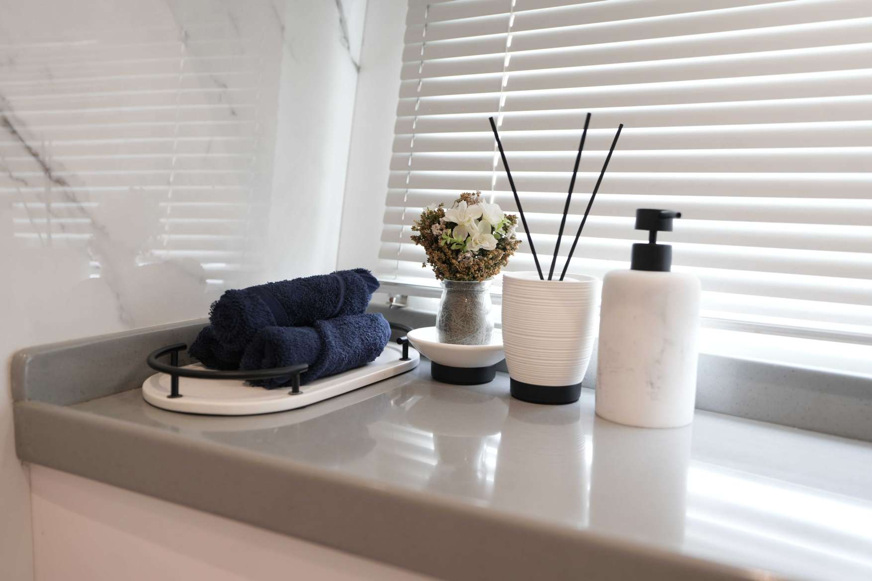 Luxury bathroom countertop on Belle 75 yacht with folded navy towels, marble soap dispenser, reed diffuser, and decorative flowers beside window blinds.