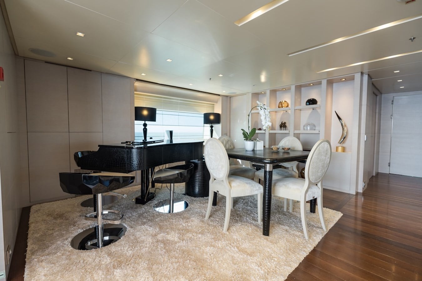 Yacht lounge with a black grand piano on a cream rug beside a dining table with white upholstered chairs