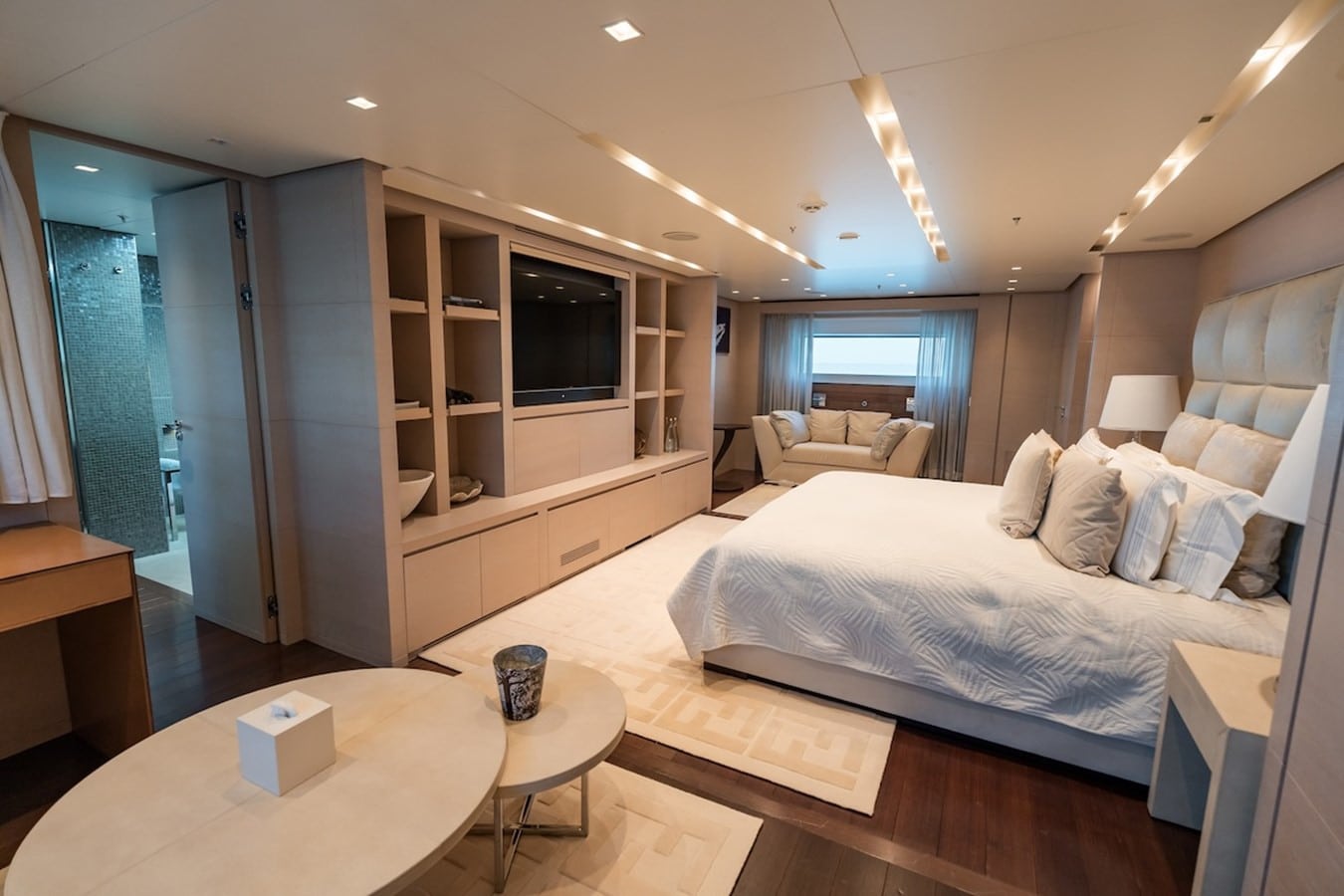 Yacht stateroom with king bed, built-in TV cabinetry, armchair by the window and en-suite bathroom beyond