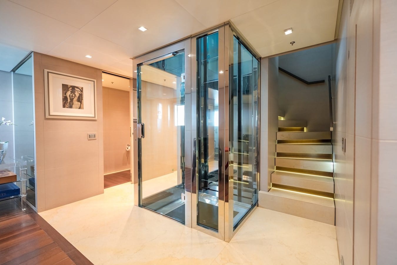 Yacht lobby with a glass and steel elevator beside an illuminated staircase and marble floor